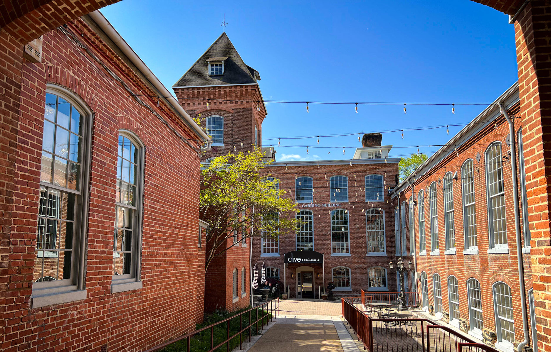 Historic brick courtyard with arched windows, string lights overhead, and a central tree. Bright blue sky enhances the warm, inviting ambiance.