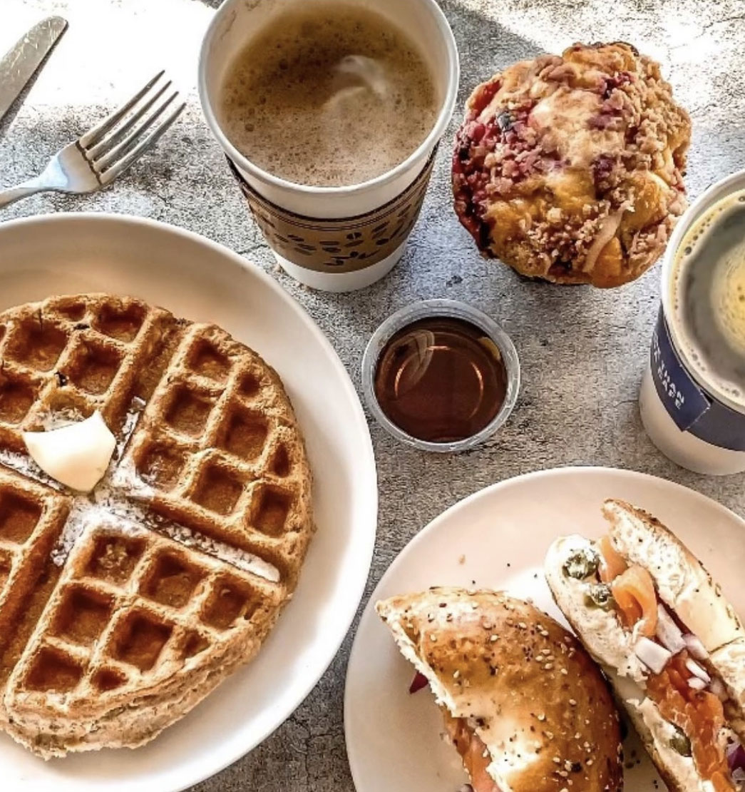 A breakfast spread with a waffle topped with butter, a bagel sandwich with salmon, a muffin, coffee cups, and syrup on a table, conveying a cozy morning vibe.