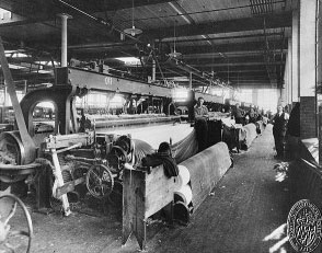 Black and white photo of a bustling textile mill with workers operating large, mechanical looms. The scene is industrial and busy, with a focus on productivity.