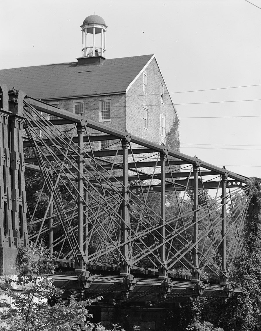 A black and white photo shows a historic iron truss bridge with intricate cross-bracing in the foreground. A large brick building with a cupola is in the background.