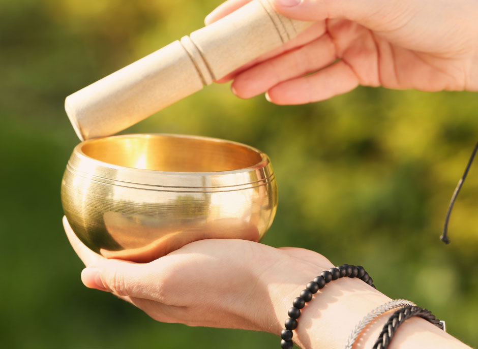 Hands holding a golden singing bowl and mallet outdoors. Sunlight highlights the bowl's smooth surface, creating a tranquil, meditative atmosphere.