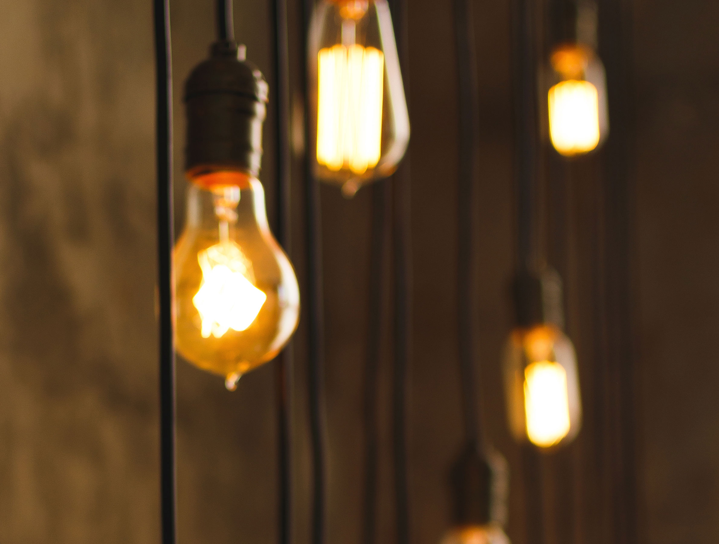 Close-up of five vintage Edison light bulbs hanging from black cords, emitting a warm, golden glow against a blurred brown background. Cozy ambiance.