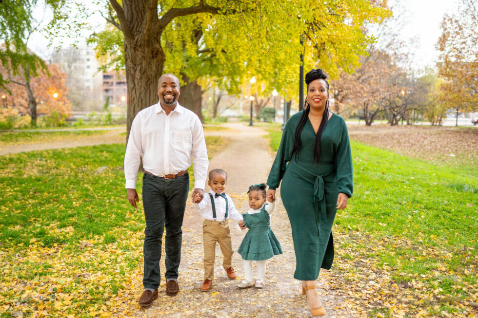 Photograph of a family of four walking through a park in autumn. The parents hold hands with two young children, dressed in coordinating green outfits, smiling warmly.