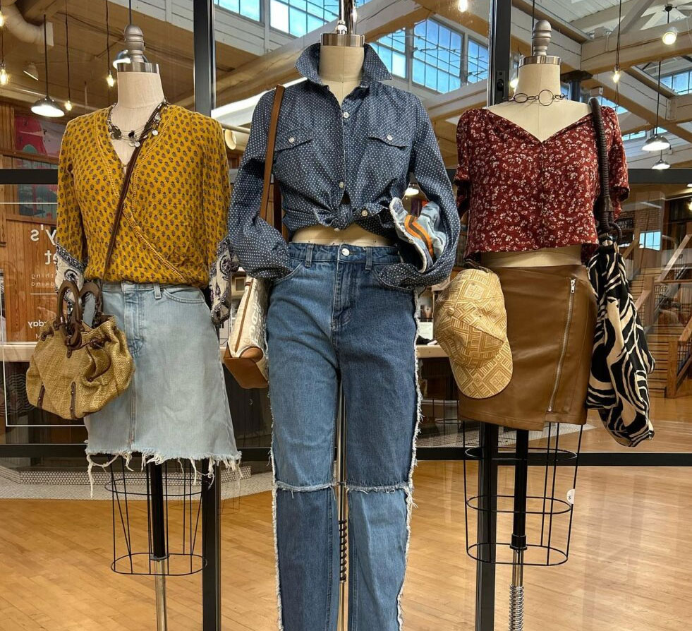 Three mannequins display trendy outfits in a boutique: a mustard blouse with denim skirt, a polka-dotted shirt with jeans, and a floral top with leather skirt. Each ensemble is accessorized with unique bags, creating a casual yet chic vibe.